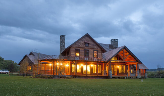 A large, rustic house with stone chimneys and a wraparound porch glows warmly with lights at dusk, set against a cloudy sky and surrounded by a grassy lawn.