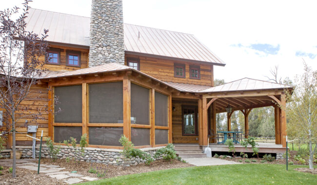 A wooden house with a stone chimney, screened porch, and covered outdoor dining area. The yard has green grass, stone pathway, and small shrubs and trees.