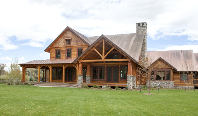 A large rustic wooden house with a steep metal roof, covered porch, stone chimney, and wide green lawn under a partly cloudy sky. The house features timber framing and stone details.