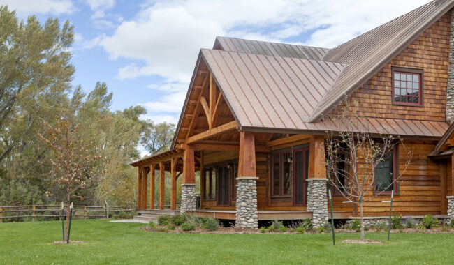 A rustic wooden house with a large porch and stone pillars stands on a well-kept lawn, surrounded by young trees and set against a backdrop of tall trees and a partly cloudy sky.