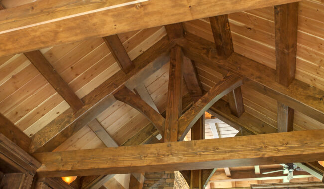 The image shows the interior ceiling of a room with exposed wooden beams and trusses, highlighting the craftsmanship of the timber framework and the warm, rustic wood paneling.