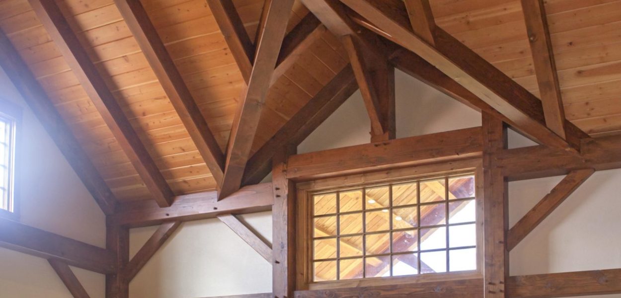 A spacious living room with high, exposed wooden beams and a vaulted ceiling. Large windows and glass doors let in natural light and reveal a green lawn outside. There is a red sofa and plants in the room.