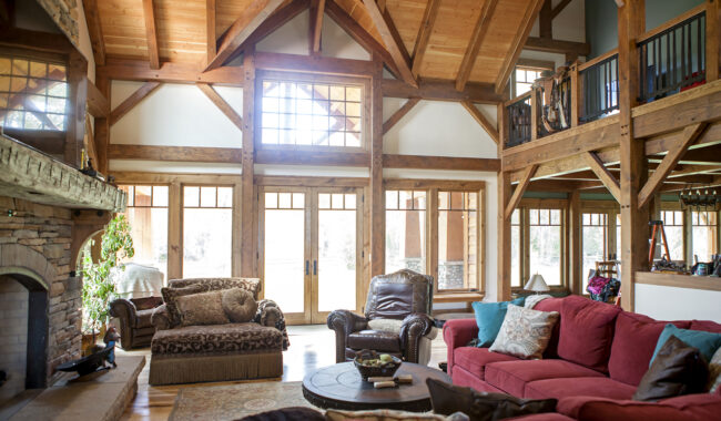 A spacious living room with exposed wooden beams, large windows, a stone fireplace, and a mix of red and brown sofas and armchairs arranged around a round coffee table on a rug.