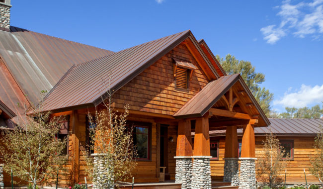 A wooden house with a brown metal roof and stone pillars sits on a well-manicured green lawn under a blue sky with scattered clouds. Young trees are planted around the house.