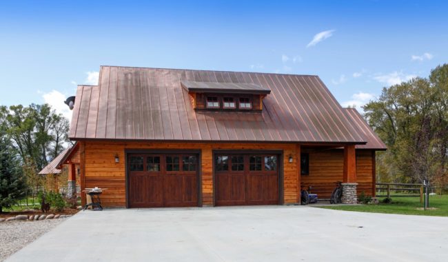 A rustic wooden house with a large copper metal roof, two wooden garage doors, stone pillars, and a wide concrete driveway, set among trees under a clear blue sky.