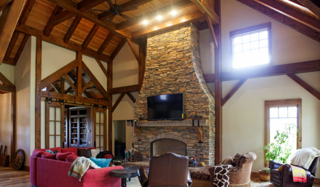 Spacious living room with exposed wooden beams, a tall stone fireplace with a TV above, large windows, cozy furniture including a red sofa, armchairs, and natural light streaming through the windows.