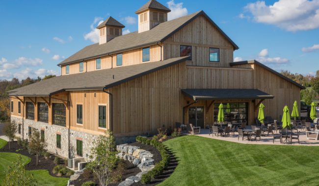 A large modern barn-style building with wood siding and stone foundation, two small cupolas on the roof, surrounded by manicured lawns and an outdoor patio with green umbrellas and tables, under a blue sky.