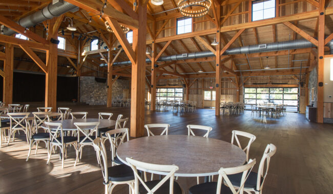 Spacious wooden event hall with high ceilings, exposed beams, and large windows. Several round tables with white chairs are arranged throughout the room. Natural light fills the space.