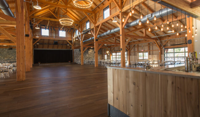 Spacious rustic event hall with exposed wooden beams, high ceilings, chandeliers, string lights, wooden floors, large windows, and a stone wall; tables and chairs are set up along the sides.