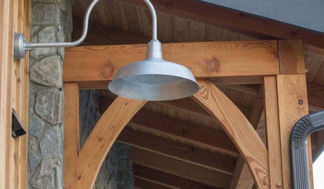 A silver metal outdoor wall light fixture is mounted on a wooden beam structure, with a stone wall and wooden ceiling visible in the background.