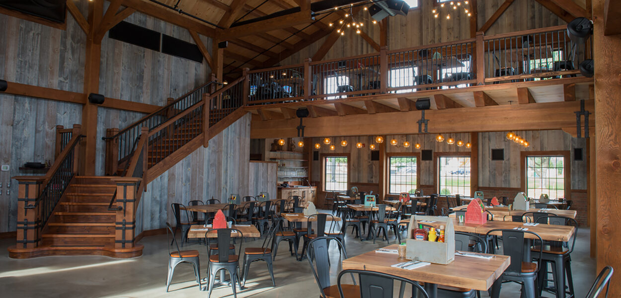 Spacious, rustic restaurant interior with wooden beams, high ceilings, modern light fixtures, and a mezzanine. Tables and black chairs are neatly arranged, and sunlight streams through large windows.