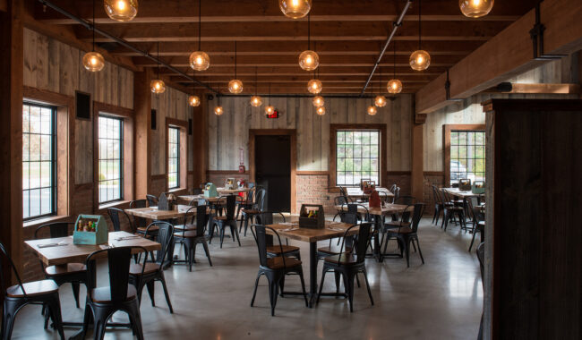 A rustic restaurant dining area with wooden beams, exposed ceilings, hanging globe lights, and several tables and chairs. Large windows let in natural light, and the room has a warm, inviting atmosphere.