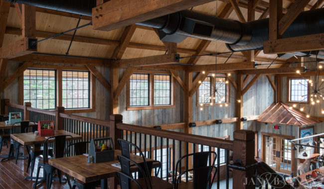 A rustic restaurant interior with exposed wooden beams, large windows, metal railings, and wooden tables with black chairs. Industrial light fixtures hang from the ceiling, and natural light fills the space.