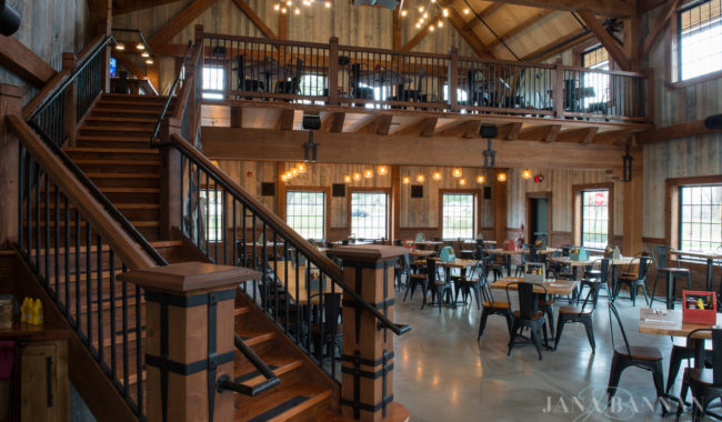 A spacious, rustic restaurant interior with wooden beams, high ceilings, a large staircase, loft seating, and metal chairs around wooden tables. Large windows let in natural light. The space feels open and inviting.