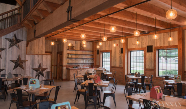 A rustic restaurant dining area with wooden walls and ceiling, hanging round light bulbs, black metal chairs, wooden tables set for guests, and large star decorations on one wall. Sunlight streams in through several windows.