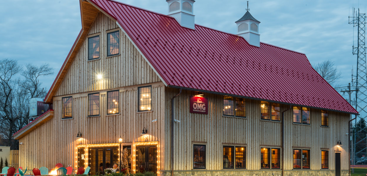 A large, rustic wooden building with a steep red metal roof, warm exterior lights, and outdoor seating. The sky is cloudy and trees without leaves are in the background. The building sign reads OMG.