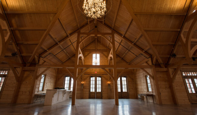 Spacious wooden hall with high vaulted ceilings, exposed beams, and a large chandelier hanging in the center. Sunlight streams through tall windows and glass doors, illuminating the polished floor. Tables line the sides of the room.