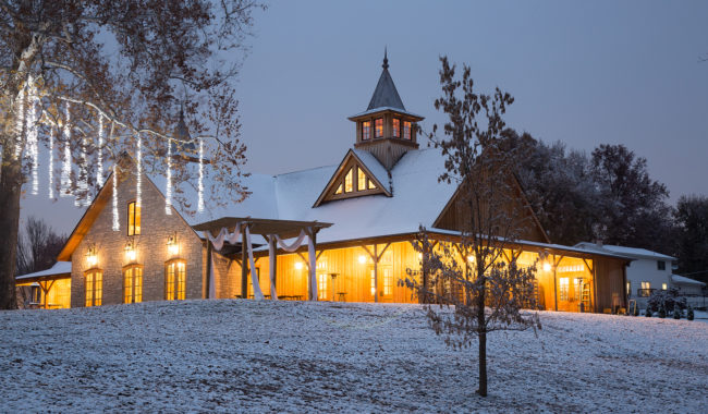 A warmly lit barn-style building with a cupola, surrounded by trees and covered in a light layer of snow at dusk. Icicle lights hang from a nearby tree, creating a cozy winter scene.