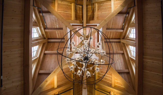 A crystal chandelier hangs from the wooden vaulted ceiling of a room, with exposed beams and several small windows letting in natural light.