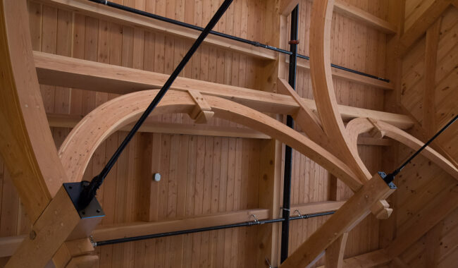 A wooden ceiling with large curved beams and black metal rods crossing diagonally, showcasing intricate timber frame construction. Warm natural wood tones and visible joints emphasize architectural detail.