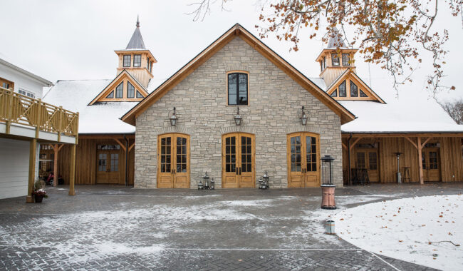 A rustic building with stone walls, wooden doors, and two towers sits in a snowy courtyard. Bare tree branches frame the scene, with light snow covering the ground and rooftops.