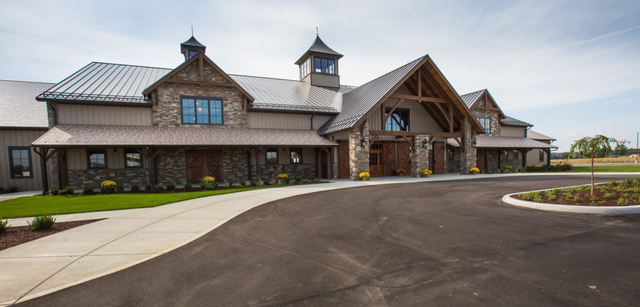 A large, rustic-style building with stone and wood accents, a peaked roof, multiple dormer windows, and a covered entrance. The driveway curves in front, surrounded by landscaped grass and flowerbeds under a blue sky.