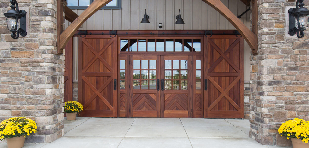 Large wooden barn-style double doors with decorative glass panels in the center, flanked by stone columns and yellow potted flowers on both sides. Black lamps are mounted on the stone columns.