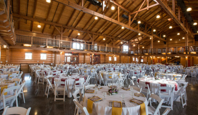 Large wooden banquet hall with high ceilings, filled with round tables covered in white cloths, set for an event. White chairs surround the tables, and small floral centerpieces decorate each table. Warm lighting fills the space.