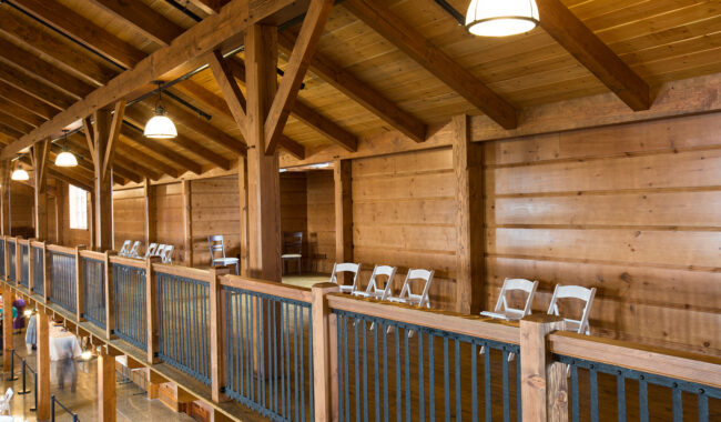 A wooden loft interior with exposed beams, overhead lights, and a row of empty white folding chairs lined up along a railing, overlooking a lower level.