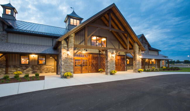 A large, rustic barn-style building with stone and wood exterior, large windows, and warm lights glowing inside, set against a cloudy evening sky with yellow flowers lining the entrance.