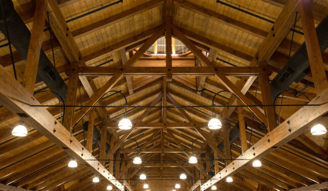 Wooden beams and trusses form the high ceiling of a large building, with numerous hanging round lights and exposed ducts visible throughout the spacious interior.