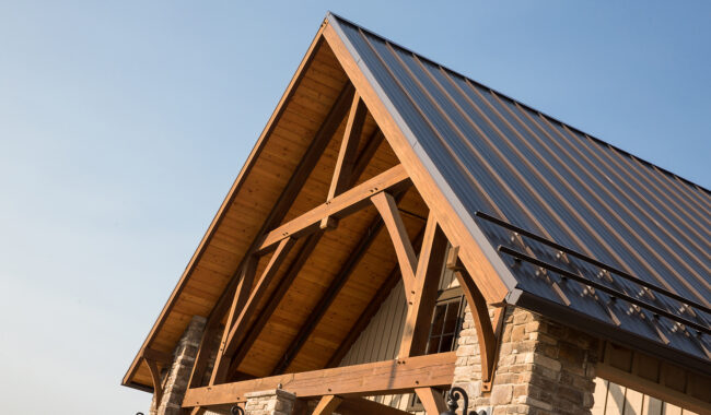 A close-up view of a house’s gable roof with exposed wooden beams and a metal roof, supported by stone columns, against a clear blue sky.