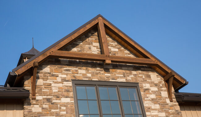 A close-up of a house exterior shows a gabled roof with exposed wooden beams, a stone facade, and a large window beneath a clear blue sky.