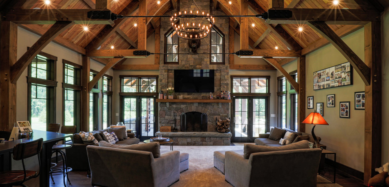 A spacious rustic living room with a high vaulted wood-beam ceiling, large windows, two gray sofas facing a stone fireplace, a central chandelier, and family photos decorating the walls.