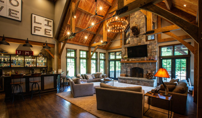 A spacious rustic living room with exposed wood beams, large windows, a stone fireplace, cozy sofas, a bar area, and warm lighting from chandeliers and lamps.