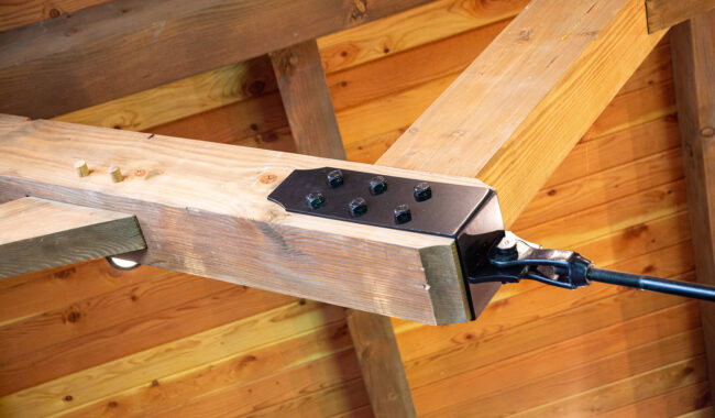 Close-up view of a wooden roof beam connection reinforced with a metal plate and bolts, attached to a black tension cable, set against a wooden ceiling with visible grain patterns.