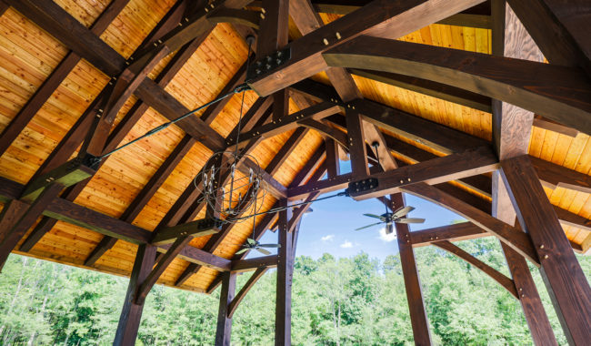 Wooden pavilion roof with exposed beams, metal hardware, two ceiling fans, and a chandelier. Sunlight highlights the natural wood, and green trees are visible outside against a blue sky.