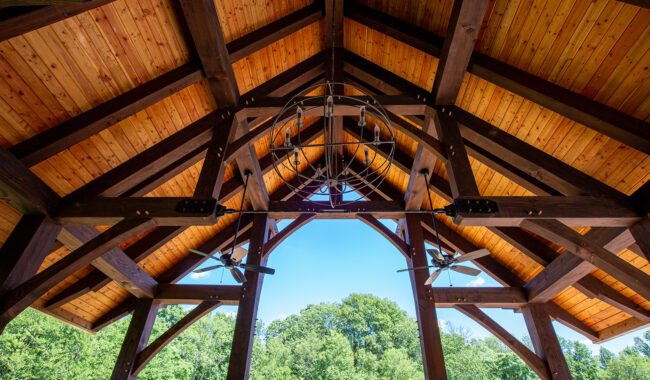View of a wooden ceiling with exposed beams, ceiling fans, and a metal chandelier, looking out to a green, leafy outdoor landscape under a clear blue sky.