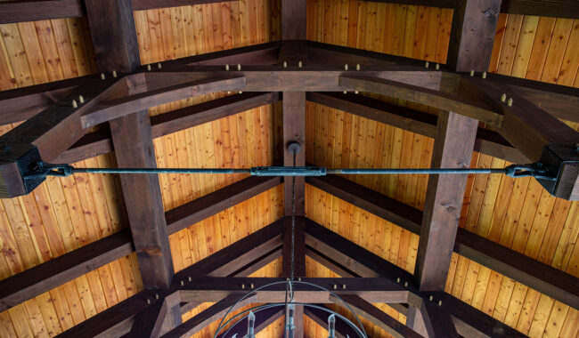 Exposed wooden beams and a tongue-and-groove wood ceiling, viewed from below, showing the architectural detail and craftsmanship of the roof structure. A metal rod spans across the beams.