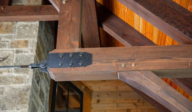 Close-up of wooden ceiling beams joined with metal hardware and bolts, next to a stone wall and wooden paneling, showcasing rustic architectural details in a building’s interior.