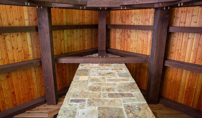 A close-up view of a wooden structure with dark wooden beams framing walls made of vertical light wood planks. A stone-tiled surface extends from the foreground to the center.