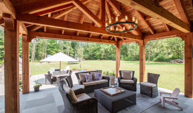 A covered outdoor patio with wicker furniture, cushions, a coffee table, and a rug. Wooden beams and a chandelier hang above. In the background, theres a large yard with trees and a white canopy tent.