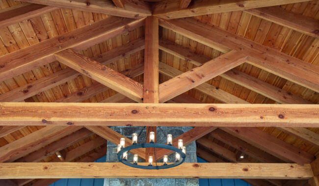 A close-up view of a wooden vaulted ceiling with exposed beams and a rustic circular chandelier hanging in the center. The wood grain is visible, giving a warm and natural look to the structure.