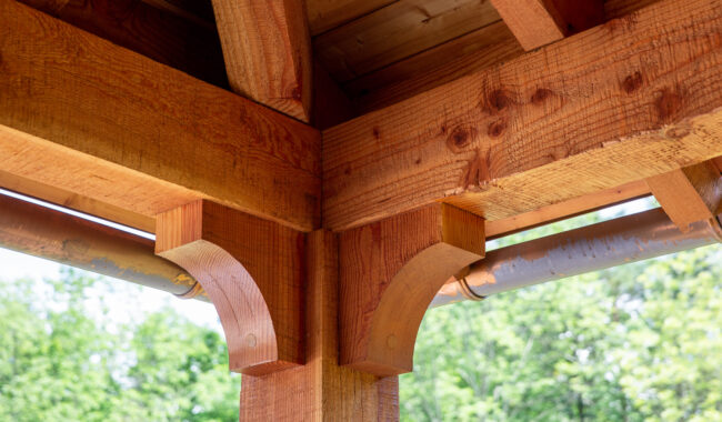 Close-up of a wooden gazebo corner showing detailed joinery and curved support brackets, with natural wood grain visible. Green trees and a clear sky are blurred in the background.