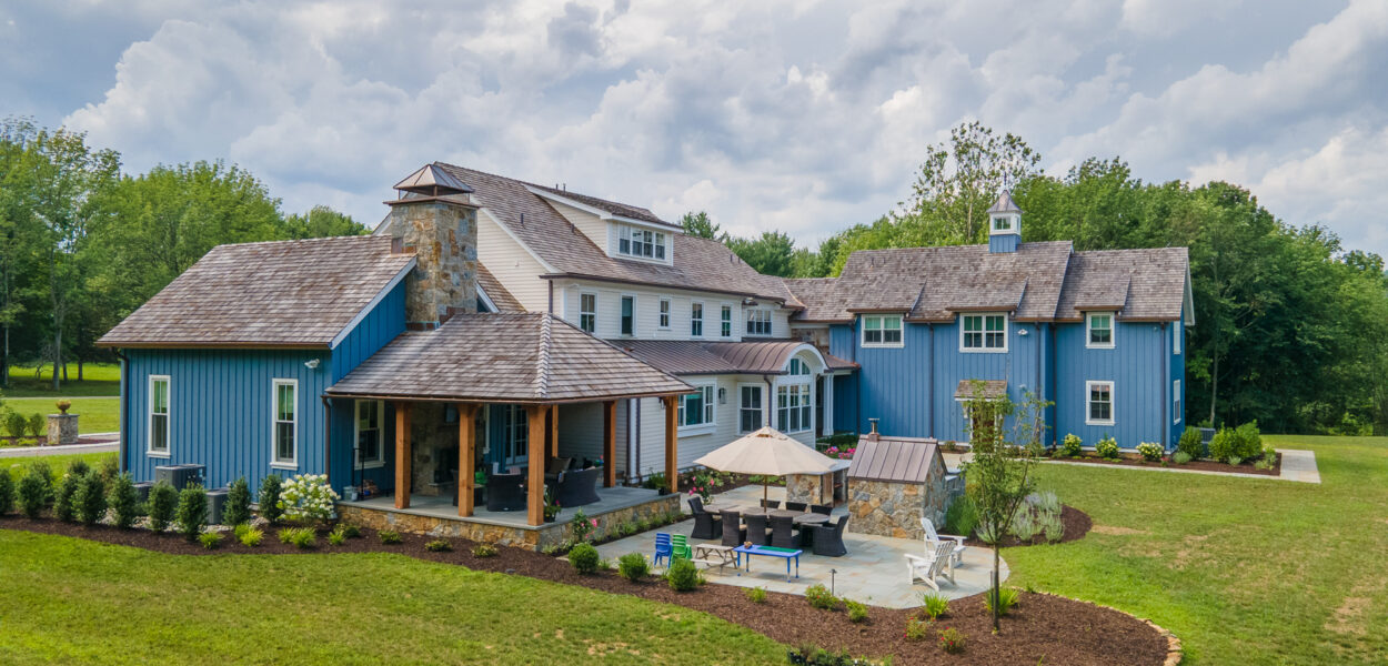 A large, blue house with wooden accents and stone features sits on a landscaped lawn. There’s a covered patio, outdoor seating area with umbrellas, and trees and shrubs surrounding the property under a cloudy sky.
