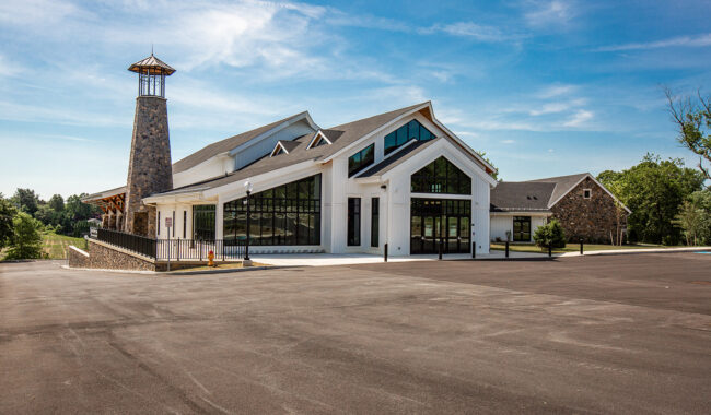 A modern white building with large glass windows and a tall stone tower stands under a blue sky. The foreground shows a spacious, empty asphalt parking lot, and trees are visible in the background.