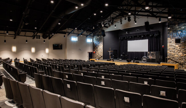 Rows of empty black chairs face a stage with instruments, equipment, and a large screen in a modern auditorium with stone walls and exposed ceiling beams.