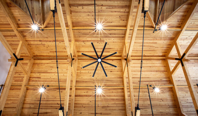 Wooden ceiling with exposed beams, a large black ceiling fan in the center, and multiple modern pendant lights hanging down, all illuminated with natural light from small skylights.