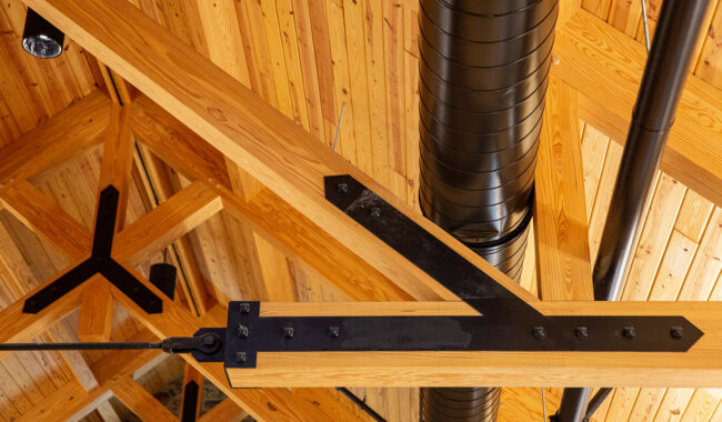 Exposed wooden beams and black metal supports on a vaulted wooden ceiling, with a large cylindrical black ventilation duct running vertically. Warm natural wood tones and industrial elements are visible.