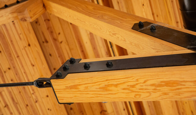 Close-up view of a wooden beam ceiling with visible black metal plates and bolts reinforcing the joints, highlighting the construction details and natural wood grain.